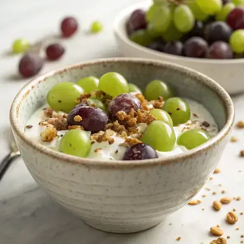 A textured bowl filled with yogurt-covered grapes and nuts, with a blurred white background and a fork resting against the bowl.