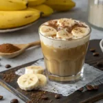 A glass of Banana Coffee topped with froth and cinnamon, surrounded by coffee beans and a banana.