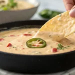 A hand dipping a tortilla chip into creamy chipotle queso in a dark cast-iron skillet, with jalapeño and red pepper specks visible.