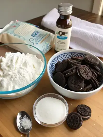 Ingredients for Oreo Fluff displayed on a kitchen table, including cream cheese, powdered sugar, vanilla extract, whipped topping, and chopped Oreo cookies.