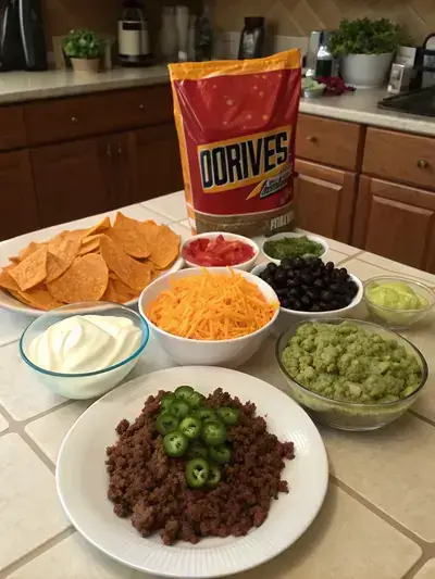 A kitchen table displaying various ingredients for loaded doritos, including Doritos, cheese, ground beef, and fresh toppings.