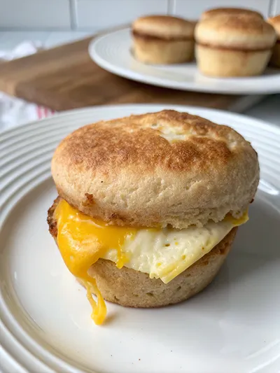 Egg and melted cheddar breakfast sandwich on an English muffin, with cheese dripping onto a white plate.