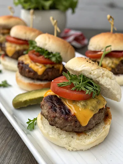 Mini beef sliders with melted cheese, tomato, and greens on small buns, served on a platter.