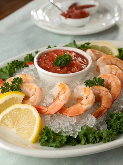 Shrimp cocktail arranged on ice with cocktail sauce, lemon wedges, and greens on a serving platter.