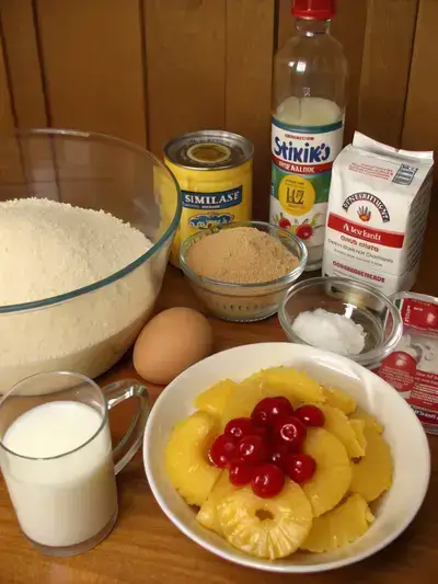 A collection of ingredients for making pineapple upside down cake, including butter, sugar, pineapple slices, cherries, flour, milk, eggs, and vanilla extract, arranged on a kitchen table.