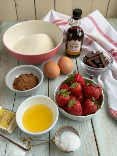 Ingredients for strawberry brownies displayed on a kitchen table, including butter, sugar, eggs, vanilla, flour, cocoa powder, salt, and fresh strawberries.